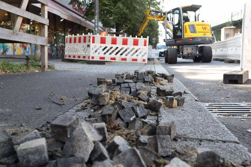 Lose Pflastersteine am Borstein im Vordergrund, schwere Maschine für die Arbeiten im Hintergrund