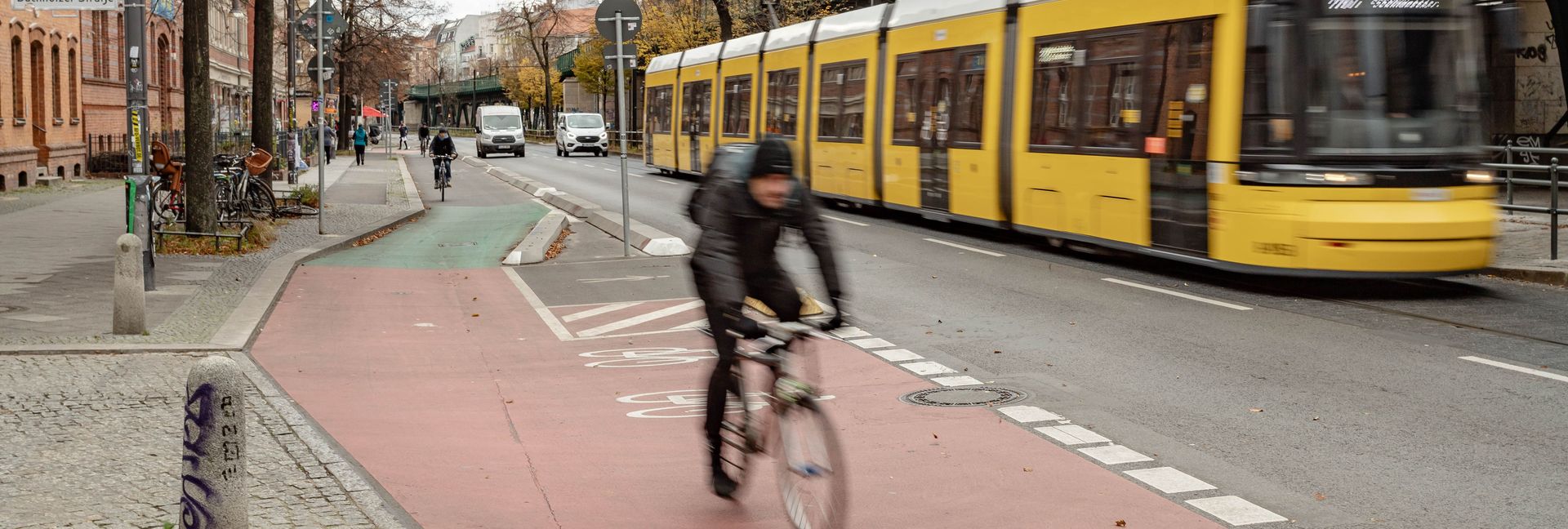 Fahrradfahrer fährt auf einem rotmarkierten Radfahrstreifen. Im Hintergrund ist das grüne Viadukt der Schönhauser Allee und eine gelbe Straßenbahn zu sehen.