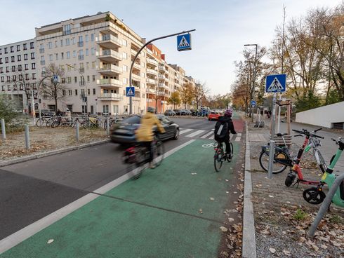 Zwei Radfahrer fahren auf einem grün beschichteten Radfahrstreifen am rechten Fahrbahnrand, vor ihnen ist ein Zebrastreifen.