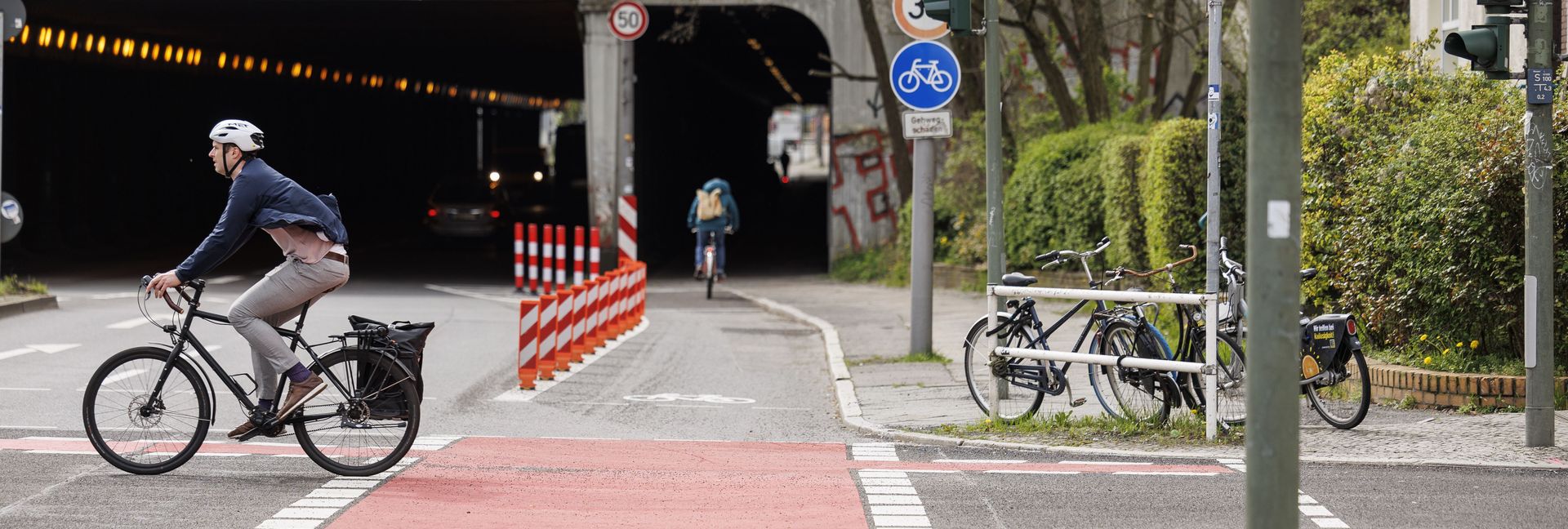 Blick auf einen Radfahrstreifen, der über eine Kreuzung führt. Im Kreuzungsbereich rot markiert, anschließend mit Pollern geschützt. Im linken Bildrand kreuzt ein Radfahrer die Straße von rechts nach links.